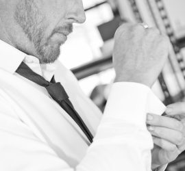 A man in a dress shirt and tie fastens his cufflink while looking down, photographed in black and white.