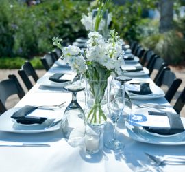 A long outdoor table set with white linens, glassware, plates, and white floral centerpieces under daylight.