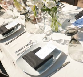 A neatly set outdoor dining table with white plates, black napkins, menus, glassware, and white flowers in vases.