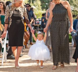A young girl in a white dress walks between two women, each holding one of her hands, at an outdoor event.