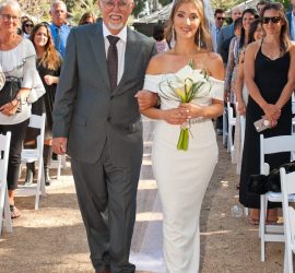 A bride in a white dress walks down the aisle holding flowers, accompanied by an older man in a suit, outdoors.
