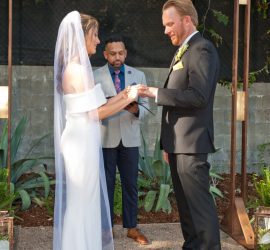 A bride and groom exchange rings during an outdoor wedding ceremony, with an officiant standing behind them.