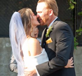 A bride and groom embrace and kiss outdoors during their wedding ceremony, both dressed in formal attire.
