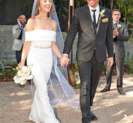 A bride and groom hold hands and smile while walking outside, with guests clapping in the background.