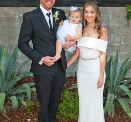 A man in a suit, a woman in a white dress, and a toddler in a white dress pose together outdoors in front of greenery.