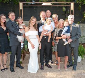 A wedding party poses outdoors, featuring a bride, groom, several adults, and young children in formal attire.