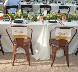 Two chairs labeled "Bride" and "Groom" at a decorated wedding reception table with place settings.