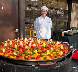 A chef in white uniform stands behind a large pan of colorful paella outdoors, with salads on a side table.