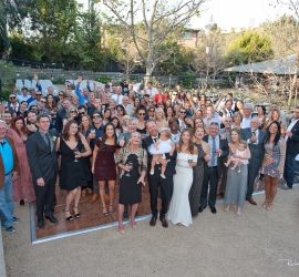 A large group of people pose together outdoors at a formal event, some holding drinks and small children.