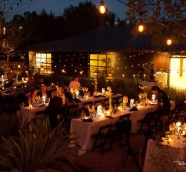 Outdoor evening event with guests seated at long tables decorated with candles and string lights, near a modern building.