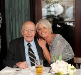 An elderly man in a suit sits at a table with a woman who is smiling and leaning close to him; flowers are on the table.