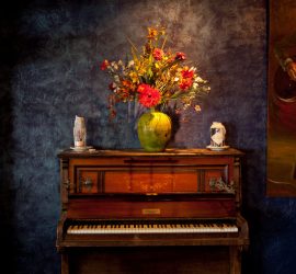 An old upright piano with a vase of flowers and two decorative skulls on top against a textured dark blue wall.