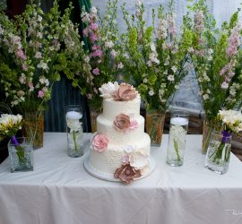 A three-tiered white cake with flower decorations on a table, surrounded by tall floral arrangements and candles.
