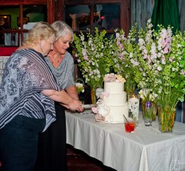 Two women stand together cutting a white tiered cake on a decorated table with flowers and drinks.