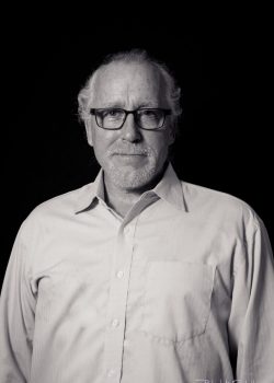 A man with glasses and a beard wears a light-colored button-up shirt, posed against a plain dark background.