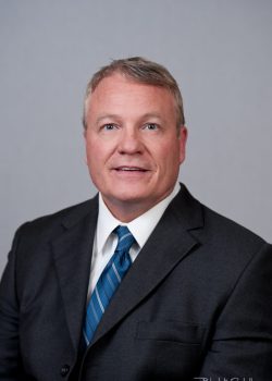 A middle-aged man in a dark suit, white shirt, and blue striped tie poses against a plain, light background.