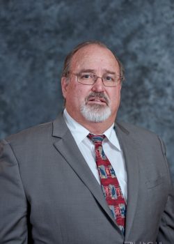 A middle-aged man in a gray suit with a patterned tie stands against a mottled gray background.
