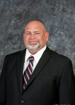A bald man with a gray goatee wearing a black suit, white shirt, and striped tie poses in front of a mottled backdrop.