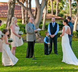 A wedding ceremony is taking place outdoors with the officiant, wedding party, and couple standing on the grass.