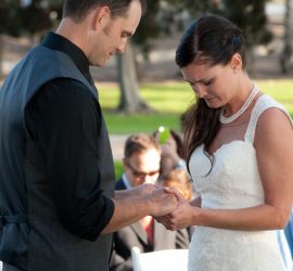 A bride and groom stand outdoors, holding hands, and looking down during their wedding ceremony.