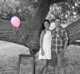 A couple stands by a tree with a pink balloon and a sign reading “Baby Manuel coming Jan 2018.”.
