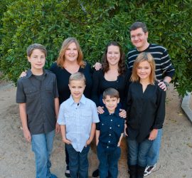 A family of seven poses outdoors in front of a green tree, with four boys, one girl, and two adults, all smiling.