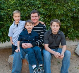 A man sits on a stone bench outdoors with three boys, all smiling, in front of a large green bush.