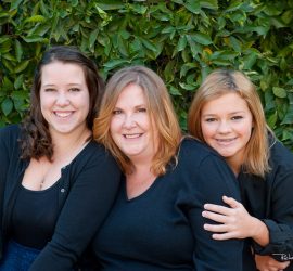 Three women sit close together in front of green foliage, all smiling at the camera and wearing dark-colored tops.