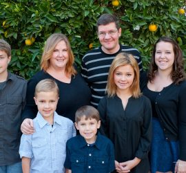 A group of seven people, including adults and children, stand together in front of a leafy orange tree.