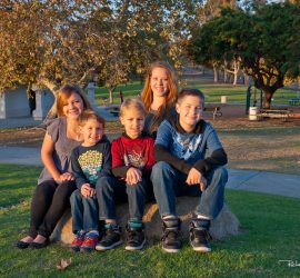 Five children sit together on a rock in a park, smiling at the camera on a sunny day.
