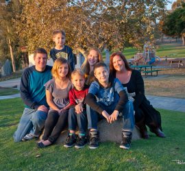 A family of seven poses together on a grassy area in a park with a playground visible in the background.