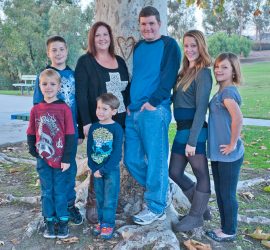 A family of seven poses in front of a large tree with carved heart in a park setting.