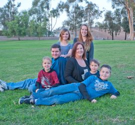 Six people, including four children and two adults, sit and kneel on grass in a park with trees in the background.