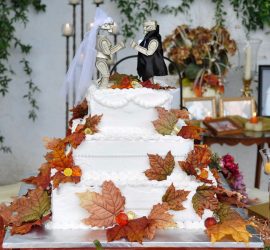 A three-tier white wedding cake with autumn leaves and two robot figurines as toppers, surrounded by candles and photos.