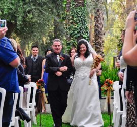 A bride in a white dress walks down an outdoor aisle on green grass, escorted by a man in a black suit.