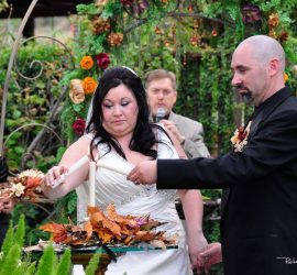Bride and groom light a unity candle during an outdoor wedding ceremony, surrounded by attendants and floral decorations.