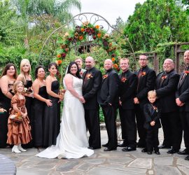 A wedding party poses outdoors under a floral arch, with the bride and groom at the center, surrounded by attendants.