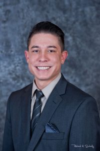 A man in a dark suit and tie smiles in front of a gray mottled background.