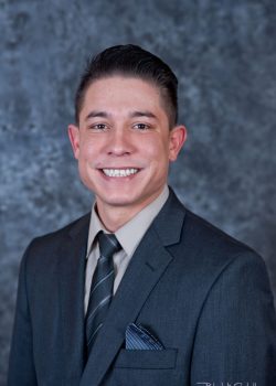 A man in a dark suit and tie smiles in front of a gray mottled background.