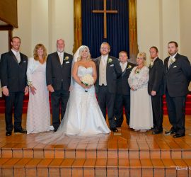 A bride and groom stand with six adults in formal attire on church steps in front of a large cross.