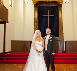 A bride and groom stand together at the front of a church, posing for a wedding photo in front of an altar and cross.