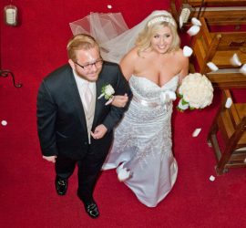 Bride and groom walk down the aisle in a church, smiling as flower petals fall around them.