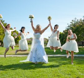 A bride and four bridesmaids in white dresses jump in the air, holding bouquets, on a grassy lawn with trees in the background.