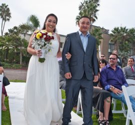 A bride in a white dress walks down the outdoor aisle with a man in a suit, guests seated on either side.
