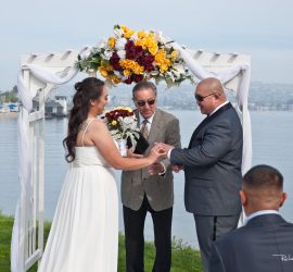 A bride and groom exchange rings under a floral arch by the water, with an officiant and a seated guest present.
