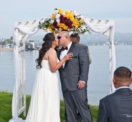 A bride and groom kiss under a floral arch by a waterfront, with guests seated and a photo display nearby.
