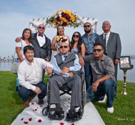 A group of people pose outdoors for a formal photo, with one man in a wheelchair at the center under a decorated arch.