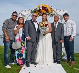 Bride and groom posing with four family members under a floral arch by a lake, with a framed photo on a stand nearby.