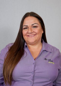 Woman with long brown hair wearing a purple "Oak Creek Energy" shirt, smiling in front of a neutral background.