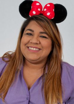 Woman wearing Minnie Mouse ears and a red polka dot bow, smiling, dressed in a purple shirt against a plain background.